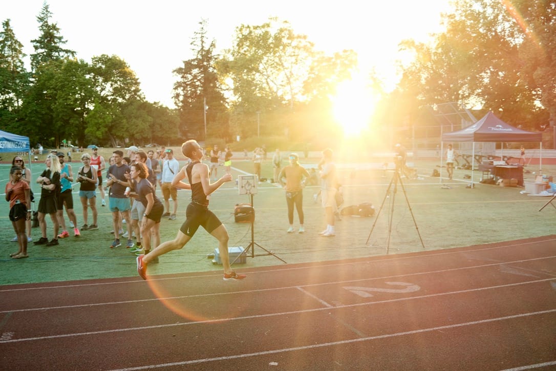 An exhausted runner crossing the finish line on a community track. A dozen people are on the turf in the center of the track. The summer sun is low and blinding, shining directly at the camera between trees