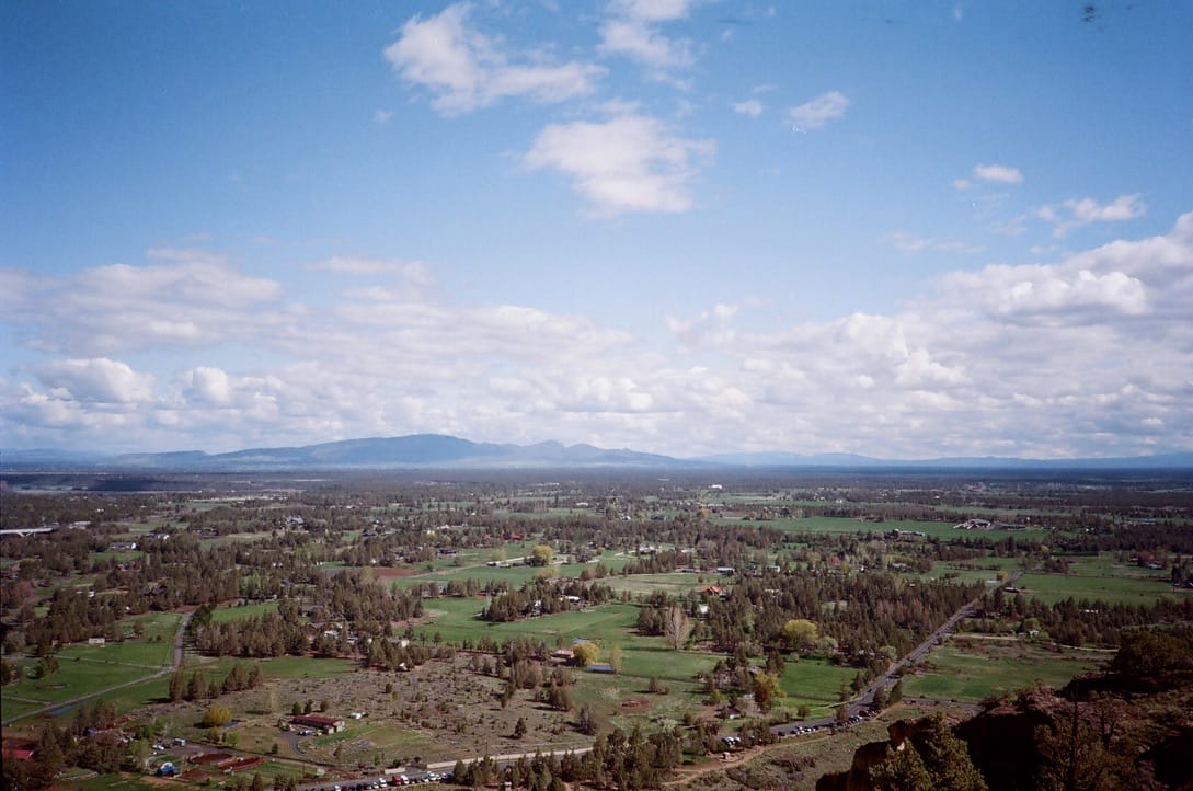 A view of farmland and evergreen trees, seen for miles from the top of a high rock. Mountains and low, white, flat clouds in the background