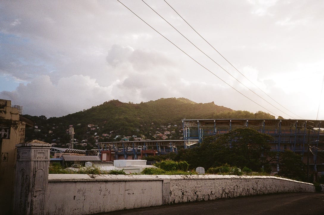  A concrete wall in the foreground, a large stadium in the middle, and a lush green mountain dotted with homes in the back. The sky is bright and cloudy, minutes after a thunderstorm